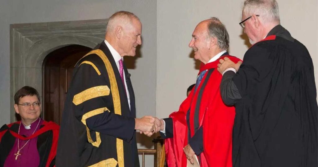 Two men in academic robes shaking hands while two other men in black clerical clothing look on