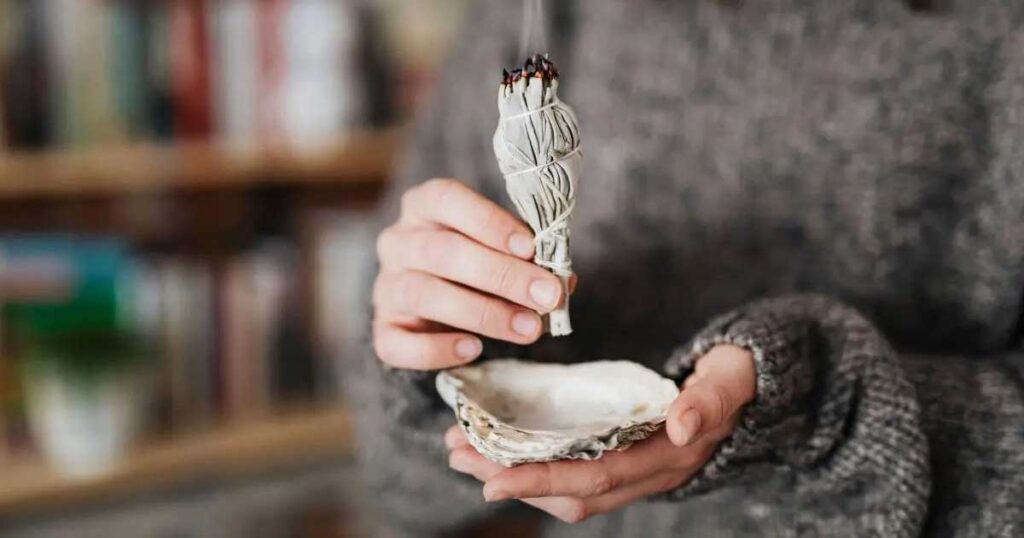 Close up of a person holding a lit burning smudging bundle over a shell to collect the ashes.